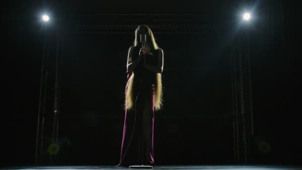 Botten bild av en chic kvinnlig solist i en lång röd klänning sjunga på en tom scen i en retro mikrofon. Artisca med långt hår ger konsert på en musikfestival i mörkret. Långsamma rörelser. — Stockvideo