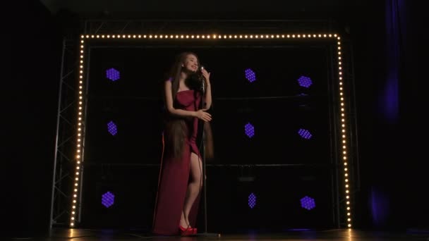 Bottom view of an attractive singer with long hair in a red dress against a background of bright lights. A woman moves sexually near a vintage microphone and sings a song emotionally. Slow motion. — Stock Video