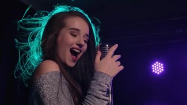 Parlak elbiseler içinde, sahnede şarkı söyleyen çekici bir şarkıcı. Kadın, arka plan dumanına ve stüdyo ışığına karşı klasik mikrofona şarkı söylüyor. Partide pop yıldızı performansı. Kapatın. Yavaş çekim.