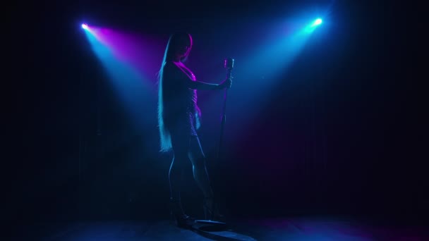 Vista inferior del vocalista pop actuando en el escenario oscuro iluminado por la luz violeta azul. Silueta oscura de mujer con el pelo largo que fluye cantando en un concierto en vivo en un micrófono vintage. Movimiento lento. — Vídeos de Stock