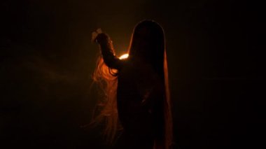 Genç ve güzel bir kadın, karanlık stüdyoda sarı ışıkla parlak sabunun arka planına doğru kıvrılır ve saçlarını sallar. Dişi poz veriyor ve yavaşça kollarını hareket ettiriyor. Siluet.
