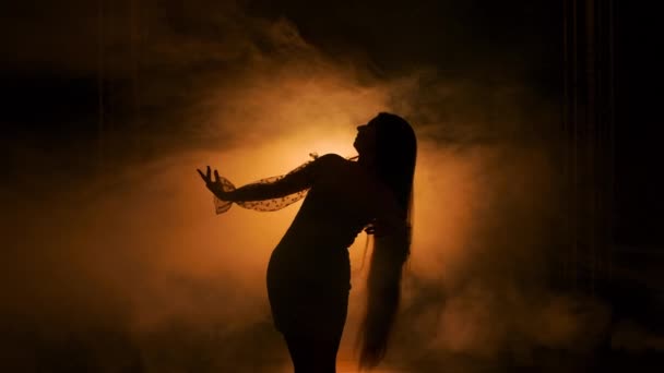 Темний силует молодої жінки, що танцює в темній димчастій студії на тлі жовтого світла. Жіночі пози в короткій сукні, виконуючи витончені чуттєві рухи руками. Близько. Повільний рух . — стокове відео