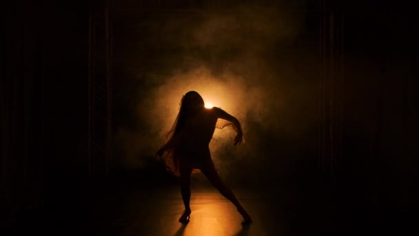 Чуттєва молода жінка танцює і розмахує своїм довгим волоссям у темній димчастій студії на тлі яскравого жовтого світла. Жінка в короткій сукні позує рухаючись у повільному русі. Силует . — стокове відео