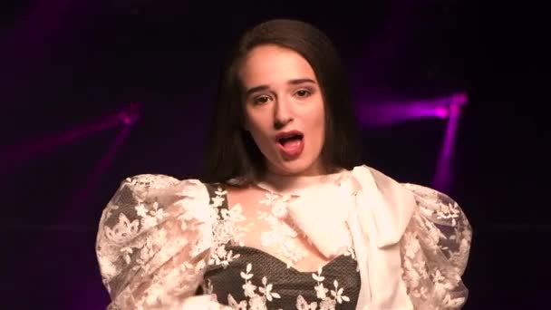 Pretty young woman in a white glamorous blouse sings in a dark studio against a background of bright purple lights. Female vocalist moves gracefully to the beat of the music in slow motion. Close up. — Stock Video