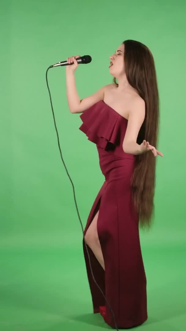 Portrait de belle jeune femme chante le chant dans le microphone et danse sur la musique. Modèle avec les cheveux longs en robe rouge profonde fente posant sur l'écran vert en studio. Vue latérale. Au ralenti. Vidéo verticale. — Video