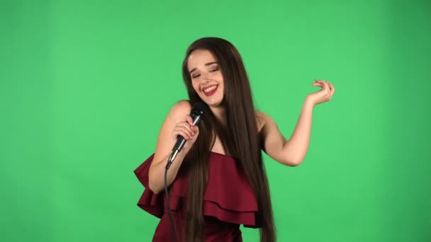 Porträt einer schönen jungen Frau, die in die Kamera blickt, Lieder ins Mikrofon singt und zur Musik tanzt. Model mit langen Haaren in rotem Kleid posiert auf grünem Bildschirm im Studio. Zeitlupe. Nahaufnahme. — Stockvideo