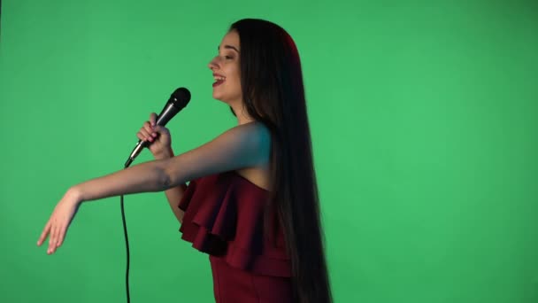 Портрет красивой молодой женщины, освещенный синим неоновым светом, поет песню в микрофон. Модель с длинными волосами в красном платье позирует на зеленом экране в студии. Вид сбоку. Медленное движение. Закрыть. — стоковое видео