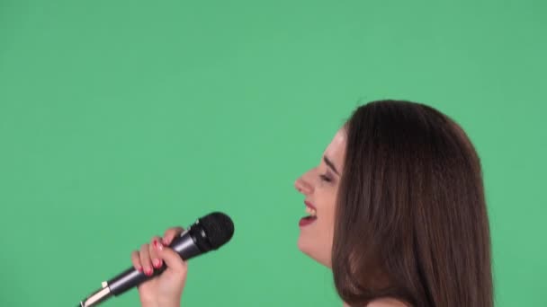 Porträt einer schönen jungen Frau singt Lied ins Mikrofon und tanzt zur Musik. Model mit langen Haaren in rotem Kleid posiert auf grünem Bildschirm im Studio. Seitenansicht. Zeitlupe. Nahaufnahme. — Stockvideo