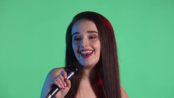 Potret wanita muda cantik diterangi oleh lampu neon merah biru menyanyikan lagu ke mikrofon. Model dengan rambut panjang dengan gaun merah berpose di layar hijau di studio. Gerakan lambat. Tutup.. — Stok Video