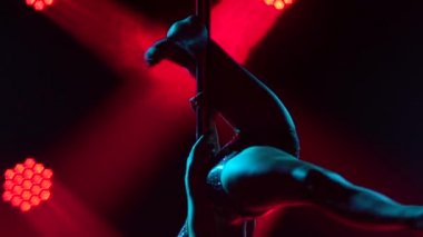 İnce, egzotik bir dansçının siluetleri direğin üzerinde güzel bir şekilde dönüyor. Parlak spor mayo giymiş genç bir kadın, karanlık bir stüdyoda kırmızı ışıkla akrobatik öğeler sergiliyor. Kapatın. Yavaş çekim.
