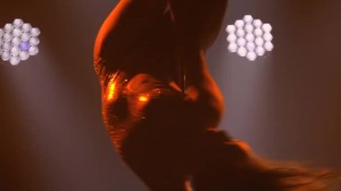 Genç dansçı striptiz direğinde dans ediyor. Kadınlar için spor konsepti. Siyah dumanlı arka planda sarı ışıklandırmalı ince, atletik bir vücudun silueti. Kapatın. Yavaş çekim.