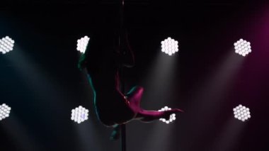 Ters dönüş, hava yürüyüşü ve amuda kalkma. Spor direği dansının akrobatik unsurları. Karanlık bir stüdyoda parlak ışıkları olan esnek vücut silueti olan genç bir kadın. Kapat..