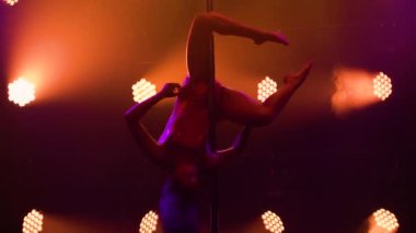 Genç bir kadın sırıkta döner, ikiye bölünür, vücudunun esnekliğini ve esnekliğini gösterir. Atletik jimnastikçi karanlık stüdyoda renkli ışıkların önünde dans ediyor. Siluet. Kapat..