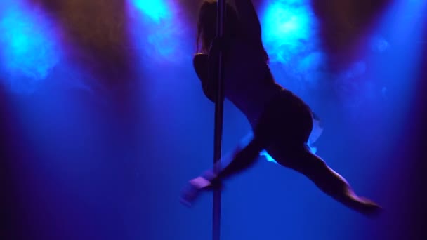 Wanita atletik yang fleksibel berputar di tiang di studio gelap dengan lampu neon biru. Menari elemen koreografi terbang, aerobik, kebugaran. Tutup.. — Stok Video