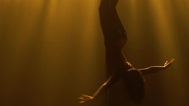 Junge Tänzerin turnt Pole Dance und führt Tricks an der Stange vor. Fitness-Konzept für Frauen. Silhouette eines schlanken athletischen Körpers auf schwarzem, rauchigen Studiohintergrund mit gelbem Licht. Nahaufnahme. — Stockvideo
