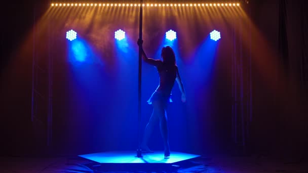 Mujer atlética flexible gira en un poste en un estudio oscuro con luces de neón azul. Bailando elementos de coreografía voladora, aeróbic, fitness. Movimiento lento. — Vídeos de Stock