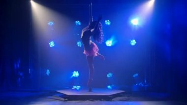 Mavi neon arka planda genç ve çekici bir kadın direğin üzerinde dans ediyor ve sonra bir öpücük yolluyor. Direk dansı spor unsurları, fitness, yaşam tarzı. Siluet. Yavaş çekim.