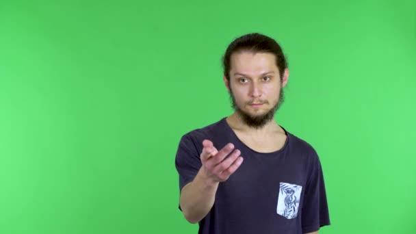 A brunet man stands and looks at the camera, beckons with a finger and waves his hand, saying hello. A man with a beard in a black T-shirt poses on a green screen in the studio. Close up. — Stock Video