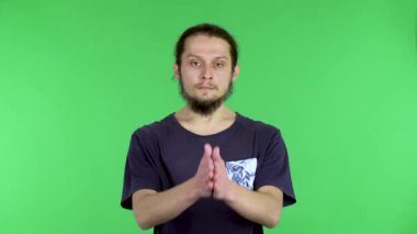 Siyah tişörtlü koyu saçlı bir genç dua jesti olarak avuçlarını bir arada tutuyor. Stüdyoda yeşil ekranda bir adam var. Kapat..