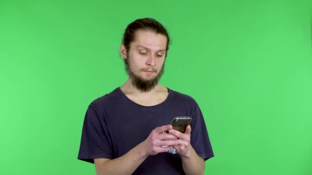 O homem lê algo em seu telefone, o que o choca, e escreve uma mensagem de texto em seu telefone. Um homem de cabelos escuros com uma barba em uma camiseta preta no estúdio em uma tela verde. Fechar. — Vídeo de Stock