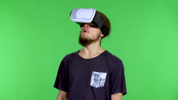 Retrato de un joven con un casco de realidad virtual o gafas 3D en la cabeza. Un moreno con barba y una camiseta negra en el estudio en una pantalla verde. De cerca.. — Vídeo de stock