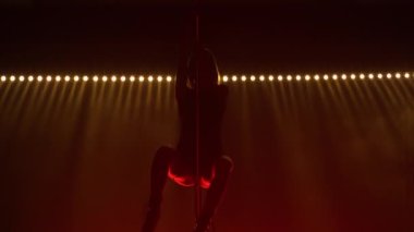 Sıska striptizci bir direkte numaralar yapar. Sarışın bir direğe tırmanıyor, havada bir yarık açıyor ve güzelce yere düşüyor. Renkli ışıkların arka planına karşı seksi bir vücudun silueti. Kapat..