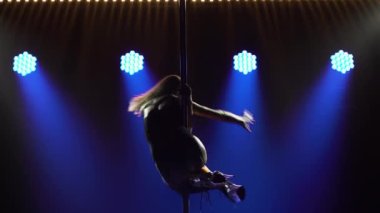 Uzun bacaklı sarışın, kollarını kendine dolamış bir direğin üzerinde dönüyor. Güzel direk dansı, kadınlar için fitness, karanlık bir stüdyoda parlak mavi ışıklar arasında striptiz gösterisi. Siluet. Kapat..