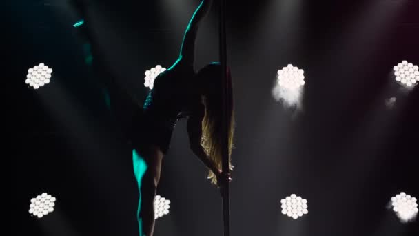 Professional pole dancer performs tricks while spinning on pole, splitting and walking in air. Stripper with long hair erotically moves on pole in dark studio with bright lights. Silhouette. Close up. — Stock Video