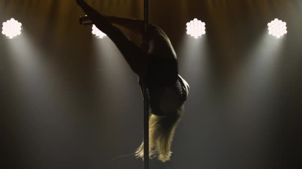 Professionelle Pole-Tänzerin führt Tricks beim Drehen an der Stange vor. Stripperin mit langen Haaren bewegt sich erotisch auf einer Stange in einem dunklen Studio mit hellen Lichtern. Erotischer Tanz, Stripshow. Silhouette. Nahaufnahme. — Stockvideo