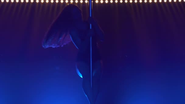 Attractive striptease dancer performs tricks on a pole. Silhouette of a sexy brunette in lingerie and high heels. Exotic dance in a dark studio with smoke and blue lights. Close up. — Stock Video
