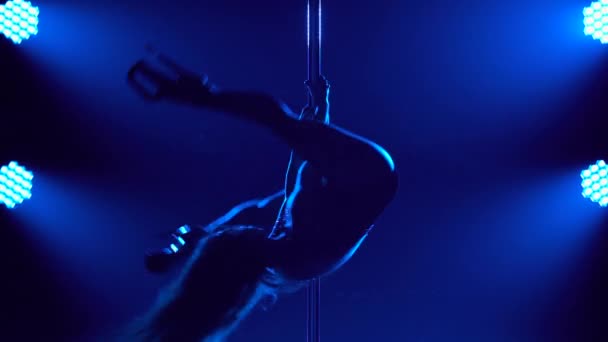 Attraktive Striptease-Tänzerin führt Tricks an einer Stange vor. Silhouette einer sexy Brünette in Dessous und High Heels. Exotischer Tanz in einem dunklen Studio mit Rauch und blauem Licht. Nahaufnahme. — Stockvideo