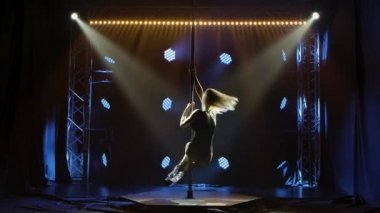 Çekici bir sarışın direkte dans eder, striptiz yapar, bacaklarını ikiye ayırır ve kalçalarını oynatır. Dinamik mavi ışıkları olan karanlık bir stüdyoda erotik direk dansı. Siluet. Yavaş çekim.
