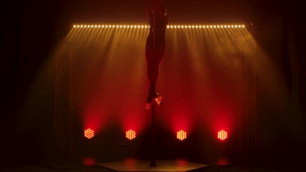 Slim stripper performs tricks on a pole. The blonde climbs a pole, performs a split in the air and falls down beautifully. Silhouette of a sexy body against a background of colored lights. Slow motion — Stock Video