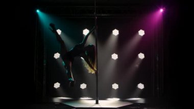 Profesyonel direk dansçısı bir direğin üzerinde dönerken, yarılırken ve havada yürürken numaralar yapar. Striptizci, karanlık bir stüdyoda, parlak ışıkları olan bir direğin üzerinde erotik hareketler yapar. Siluet. Yavaş çekim.