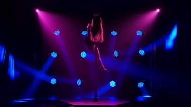 Elmas elmasları ve yüksek topuklu ayakkabıları olan mayolu seksi esmer. Striptiz dansçısı karanlık stüdyoda esneme ve esneklik gösteriyor mavi mor spot ışıklarının arka planına karşı. Yavaş çekim