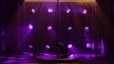 Striptiz dansçısı direğin yanında yerde yatarken cinsel olarak hareket eder. Direk dansı, fitness ve spor. Genç bir kadının atletik fiziğinin silüeti. Mor arka plan. Kapatın. Yavaş çekim.