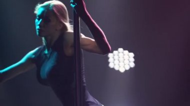 Seksi sarışın bir direğin üzerinde dans eder ve siyah iç çamaşırları ve yüksek topuklularla üzerinde döner. Erotik dans. Duman ve neon ışıkla karanlık bir stüdyoda çekildi. Parlak neon ışık efektleri. Kapatın. Yavaş çekim.