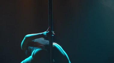 Striptiz yapan seksi zayıf kadın ve gece kulübünde bir direğin üzerinde dönen. Güzel sarışın striptizci, neon ışıkla sahnede erotik hareketler yapıyor. Siluet. Kapatın. Yavaş çekim.