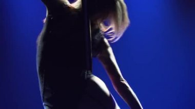 Uzun bacaklı sarışın, kollarını kendine dolamış bir direğin üzerinde dönüyor. Güzel direk dansı, kadınlar için fitness, karanlık bir stüdyoda parlak mavi ışıklar arasında striptiz gösterisi. Kapatın. Yavaş çekim.