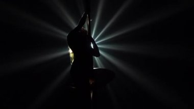 Direğin yanında dans eden bir striptizcinin siyah siluetinin alt görüntüsü. Bir direk dansçısı karanlıkta, dönen ışık huzmelerine karşı erotik bir şekilde hareket eder. Kapatın. Yavaş çekim.