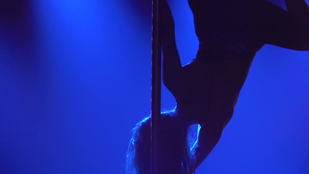 Ελκυστική χορεύτρια στριπτίζ εκτελεί κόλπα σε ένα πόλο. Η σιλουέτα μιας σέξι μελαχρινής με στενό κουστούμι και ψηλά τακούνια. Εξωτικός χορός σε σκοτεινό στούντιο με καπνό και μπλε φώτα. Κλείσε. Αργή κίνηση. — Αρχείο Βίντεο