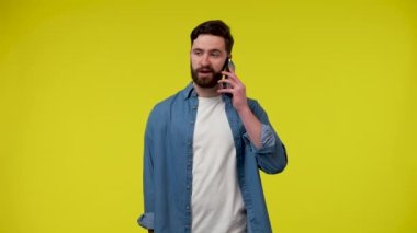 Cep telefonuyla konuşan ve bir şeye şaşıran bir adamın portresi. Stüdyoda sarı arka planda beyaz tişörtlü ve mavi kot gömlekli bir adam var. Kapatın. Yavaş çekim hazır 59.97fps.