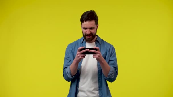 Игрок, использующий смартфон, играет в мобильное игровое приложение. Красивый мужчина в белой футболке и синей джинсовой рубашке в студии на жёлтом фоне. Закрывай. Готовность к медленному движению 59,97fps. — стоковое видео