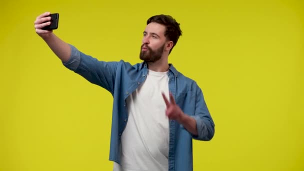 Muž, který si bere do telefonu selfie, pózuje a dělá vítězný gesto. Muž v bílém tričku a modré džínové košili ve studiu na žlutém pozadí. Zavřít. Zpomalení připraveno 59.97fps. — Stock video