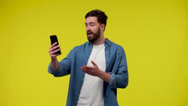 Портрет чоловіка, який робить відеодзвінок на телефон, махає рукою, вітає співрозмовника. Красивий бородатий чоловік в студії на жовтому тлі. Близько. Повільний рух готовий 59.97fps . — стокове відео