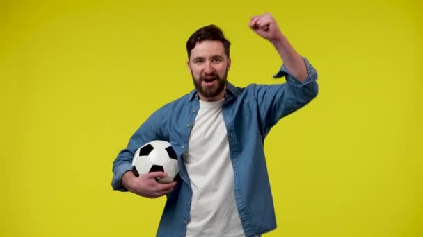 Ein stattlicher bärtiger Mann mit einem Fußballball in der Hand, der sich Sorgen macht und die Fußballmannschaft anfeuert, feiert den Sieg. Ein Mann im Studio auf gelbem Hintergrund. Aus nächster Nähe. Zeitlupe bereit 59.97fps. — Stockvideo