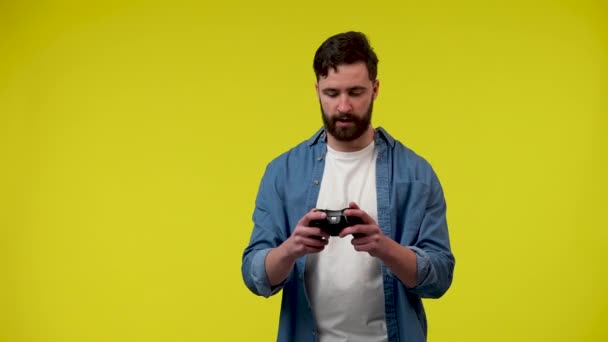 Человек, играющий в видеоигру с беспроводным контроллером, радуется победе. Мужчина в белой футболке и джинсовой рубашке в студии на жёлтом фоне. Закрывай. Готовность к медленному движению 59,97fps. — стоковое видео