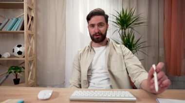 Sakallı bir adam elinde bir kalem çevirir ve masada otururken bir ekranın önünde konuşur. Video konferansı. Ev ofisindeki bir masada oturan bir adam. Kapatın. Yavaş çekim hazır 59.97fps.