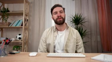 Sakallı bir adam masada otururken elini sallıyor ve ekranın önünde konuşuyor. Video konferansı. Ev ofisindeki bir masada oturan bir adam. Kapatın. Yavaş çekim hazır 59.97fps.