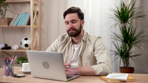 Hombre barbudo guapo utiliza el ordenador portátil mientras está sentado en la mesa en la oficina en casa. Un hombre revisa su correo electrónico en línea o trabaja en una computadora. Trabajo remoto. De cerca. Cámara lenta lista 59.97fps. — Vídeos de Stock